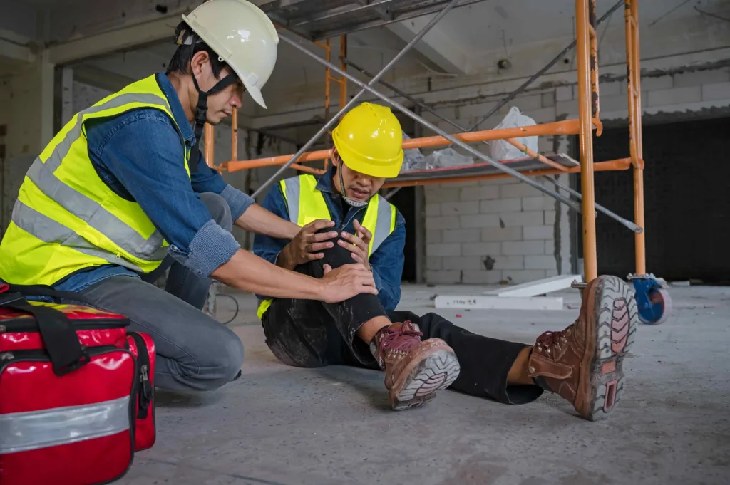 Construction worker receiving first aid for leg injury at job site, highlighting workplace accident, safety protocols, and workers’ compensation claim. Construction worker receiving first aid for leg injury at job site, highlighting workplace accident, safety protocols, and workers’ compensation claim.
