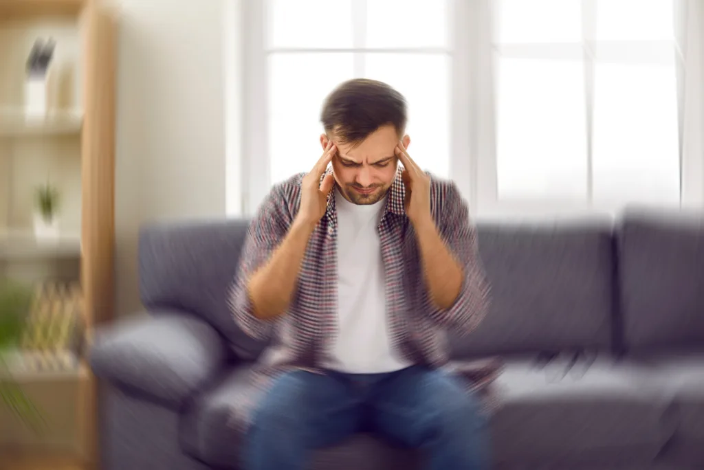 Man suddenly feels dizzy and takes a seat on the sofa. Young guy feeling pain and spinning sensation in his head. Headache, vertigo, health problem, brain tumor concept