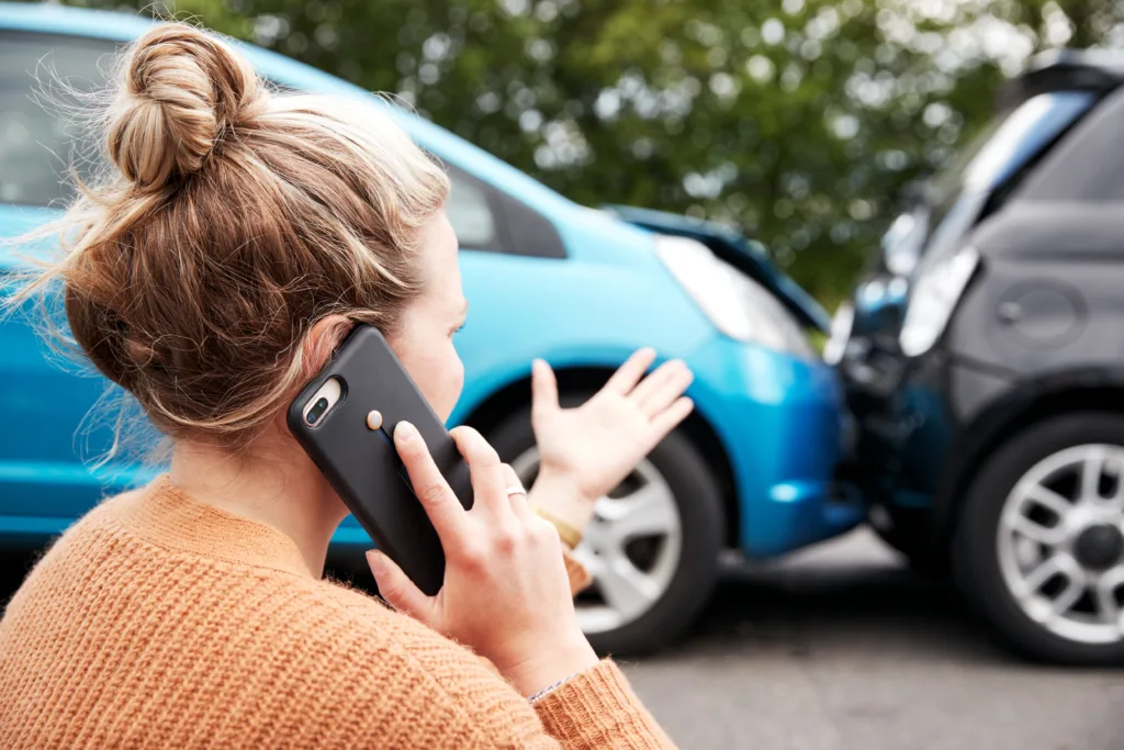 Woman calling insurance company after rear-end car accident, standing near damaged vehicles and discussing auto collision claim.