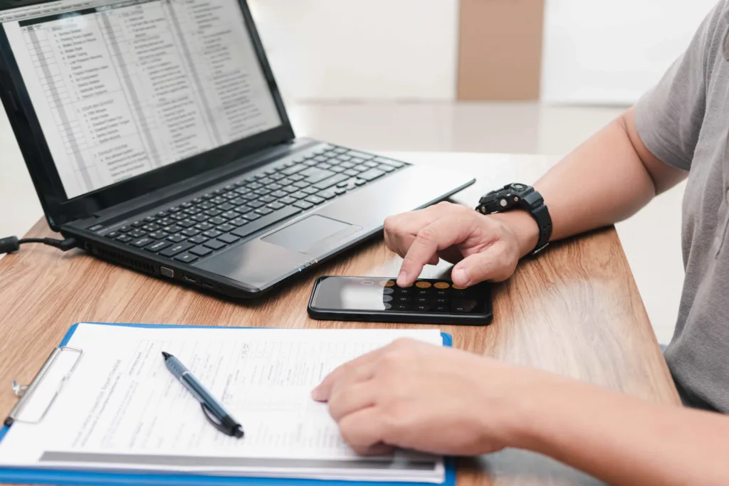 Person calculating expenses on smartphone calculator with laptop and paperwork on desk.