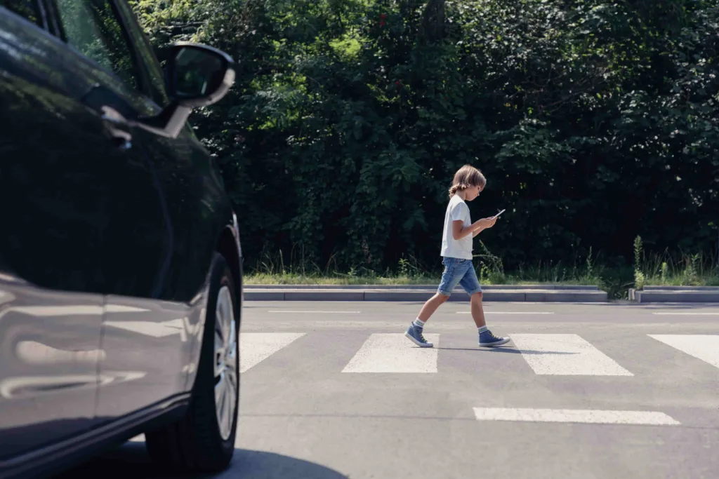 Child walking across crosswalk while looking at smartphone with car approaching in foreground.