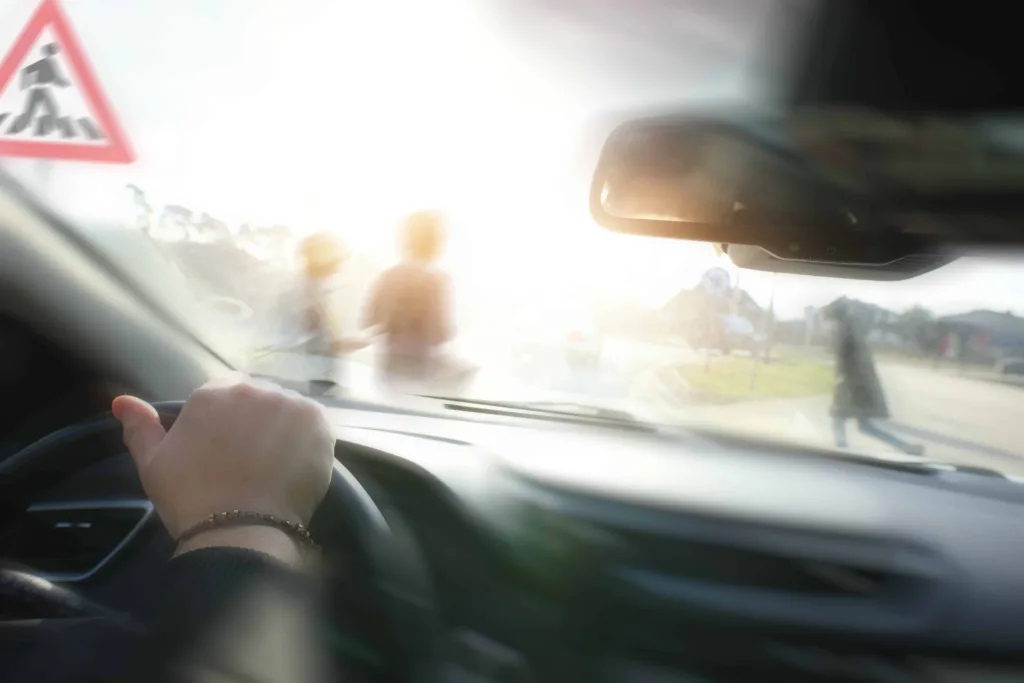 Driver view through windshield with sun glare and pedestrians crossing street near crosswalk sign.