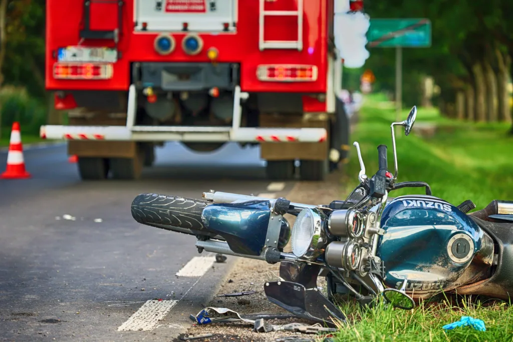 Motorcycle accident scene with a fallen Suzuki bike on the roadside and a fire truck parked in the background.